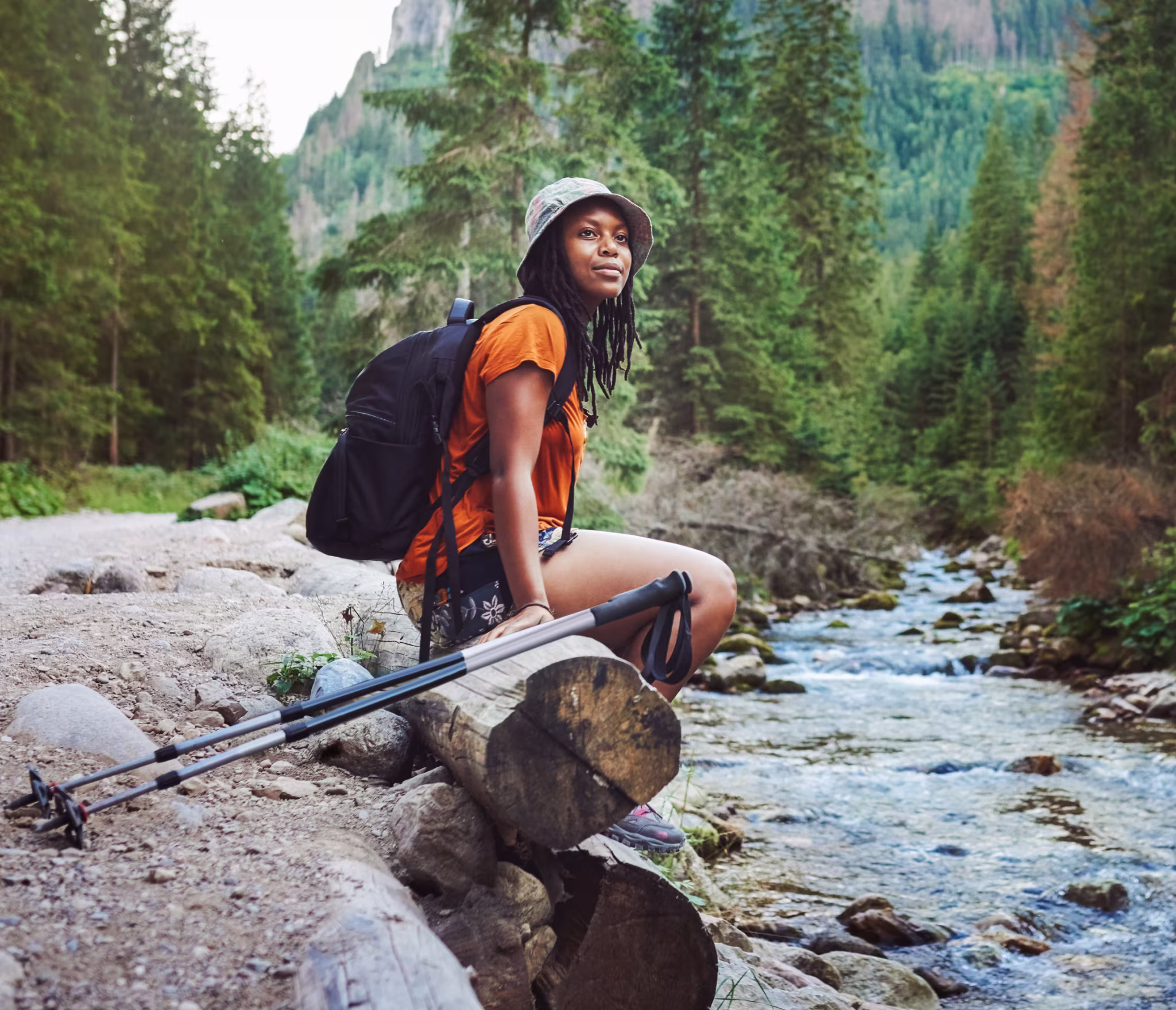 A person wearing a bucket hat, orange shirt, and backpack sits on a log beside a rocky forest stream. Trekking poles rest nearby, and tall evergreen trees fill the mountainous landscape in the background.