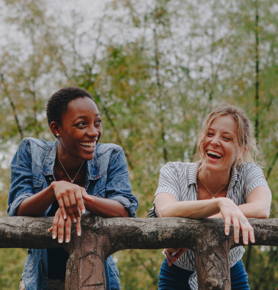 Two women laughing together while leaning on a stone railing outdoors, with trees in the background. One wears a denim jacket, and the other wears a striped shirt.