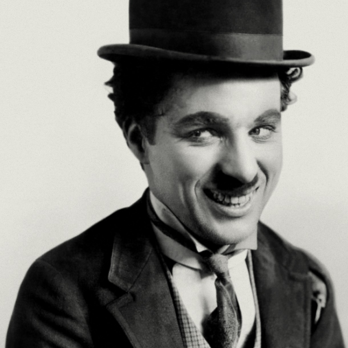 A black-and-white portrait of a smiling man wearing a bowler hat, dark suit, narrow tie, and a small toothbrush-style mustache.