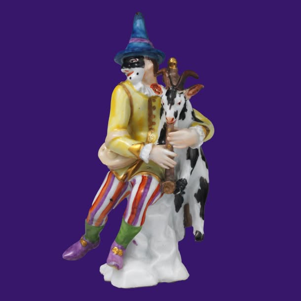 A porcelain figurine of a masked, brightly dressed performer in a pointed blue hat holding a black-and-white spotted goat against a solid purple background.