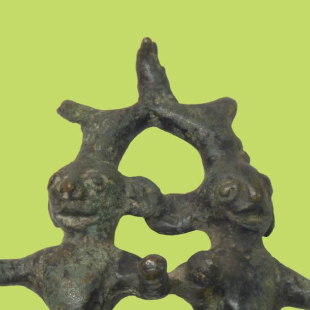 Close-up of a small dark bronze figurine showing two stylized human-like faces beneath an arched, branching form, set against a bright yellow-green background.