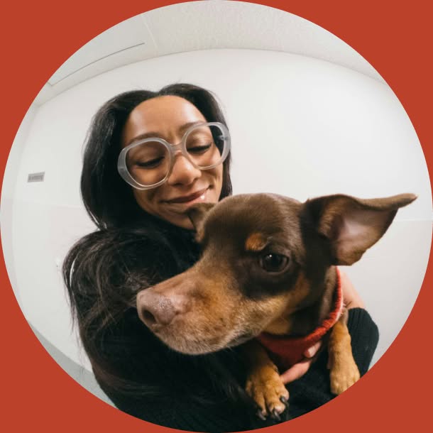 A smiling woman with long dark hair and large clear-framed glasses holds a small brown dog close to her chest, framed within a circular photo on a rust-red background.
