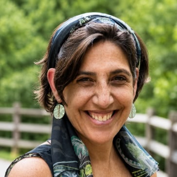 Portrait of Julia Schmidt smiling outdoors, wearing a patterned headscarf and dangling earrings against a softly blurred green background.