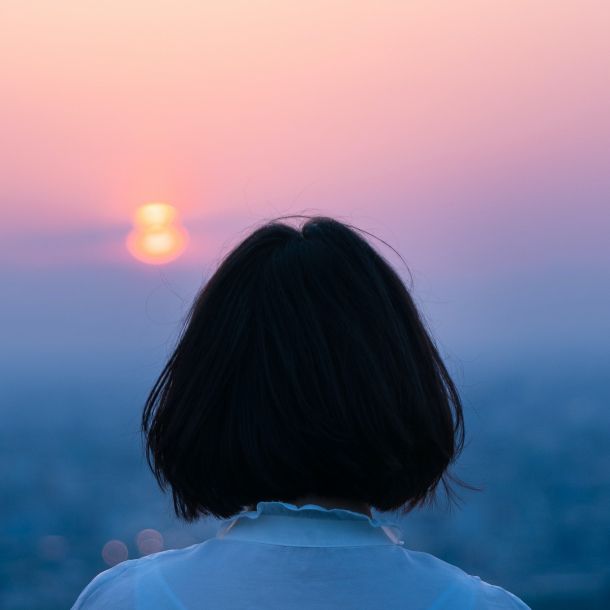 A person with short dark hair seen from behind, looking toward a glowing sunset over a hazy blue landscape.