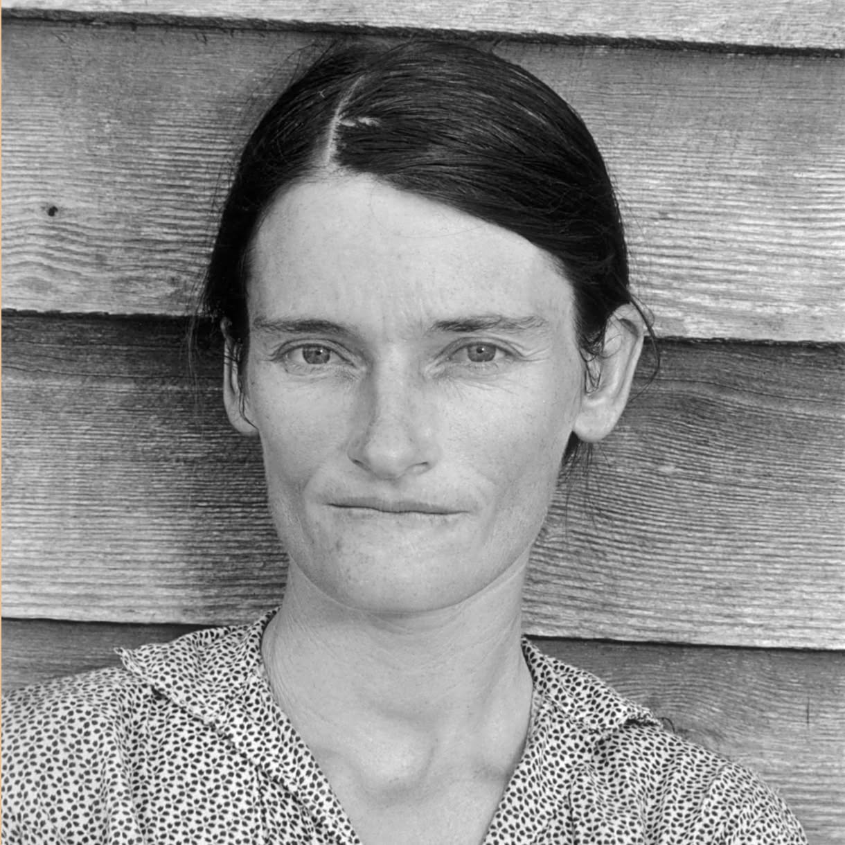 Black-and-white portrait of a woman with dark hair pulled back and parted to one side, wearing a patterned blouse. She faces the camera with a subtle, closed-mouth smile against a background of horizontal wooden siding.