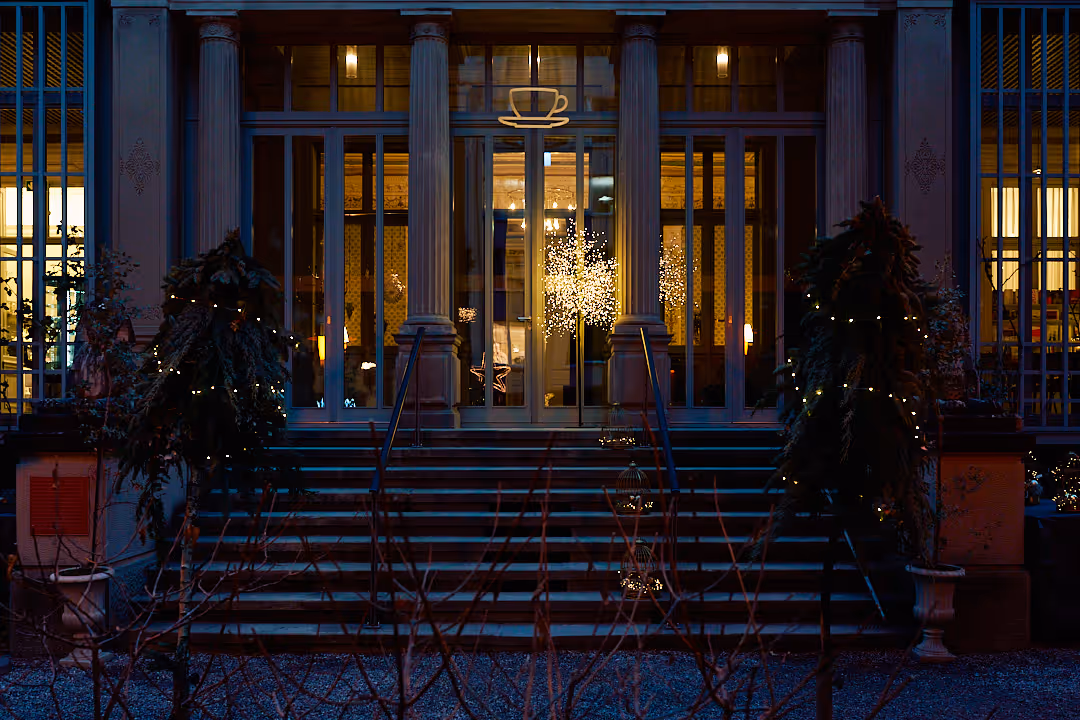 Eingang eines Cafés mit großen Glasfenstern und Treppen, beleuchtet bei Nacht mit dekorativen Lichterketten an Pflanzen.