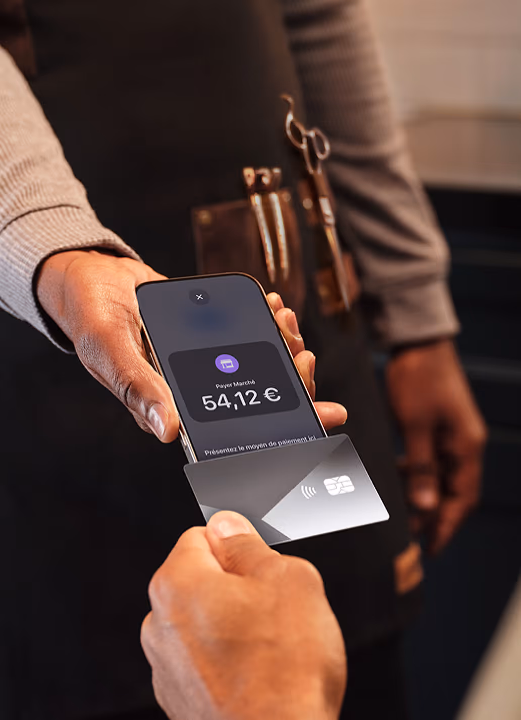 Person holding a smartphone displaying a payment of 54,12 € while another hand holds a contactless payment card near the phone.