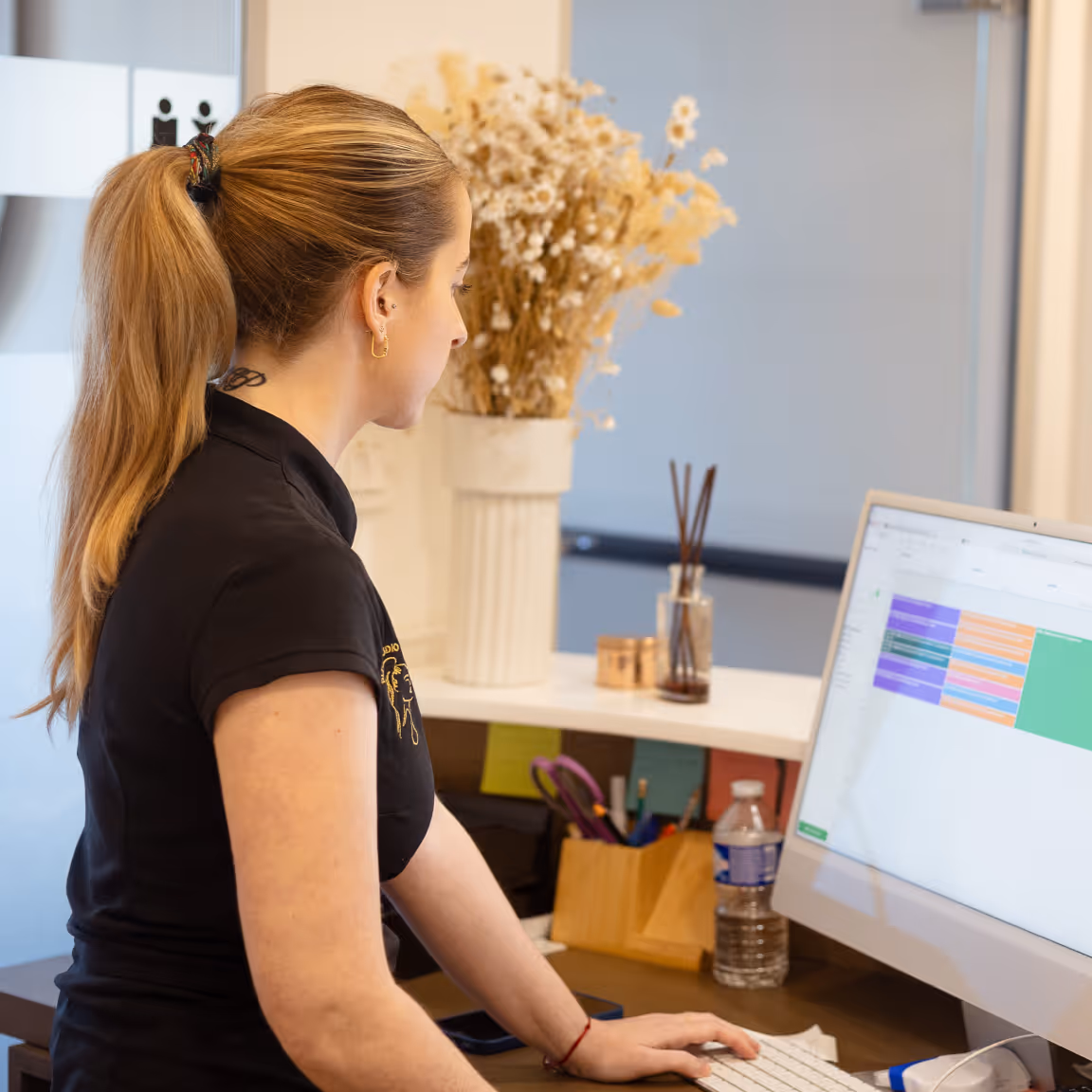 Woman with ponytail working on a desktop computer at a tidy desk with a water bottle and dried flowers.
