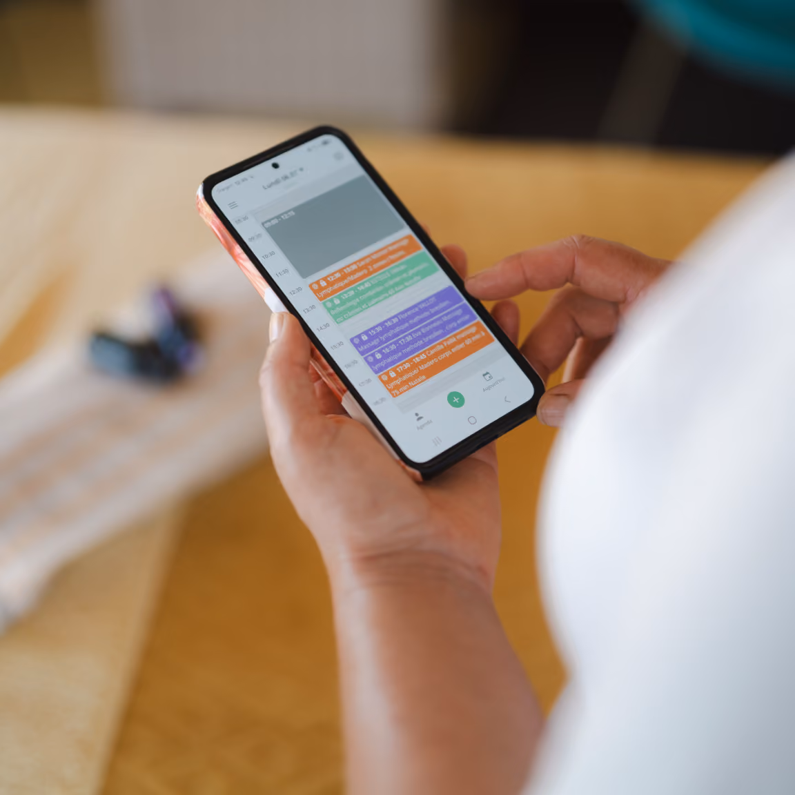 Person holding a smartphone displaying a daily schedule app with appointments color-coded in orange, green, and purple blocks for various times throughout the day.