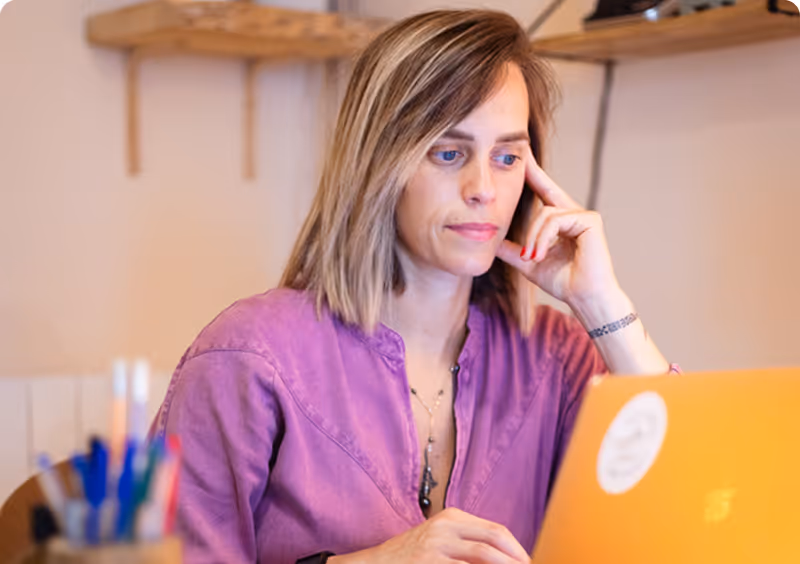 Woman with shoulder-length hair in a purple shirt working on an orange laptop at a desk.