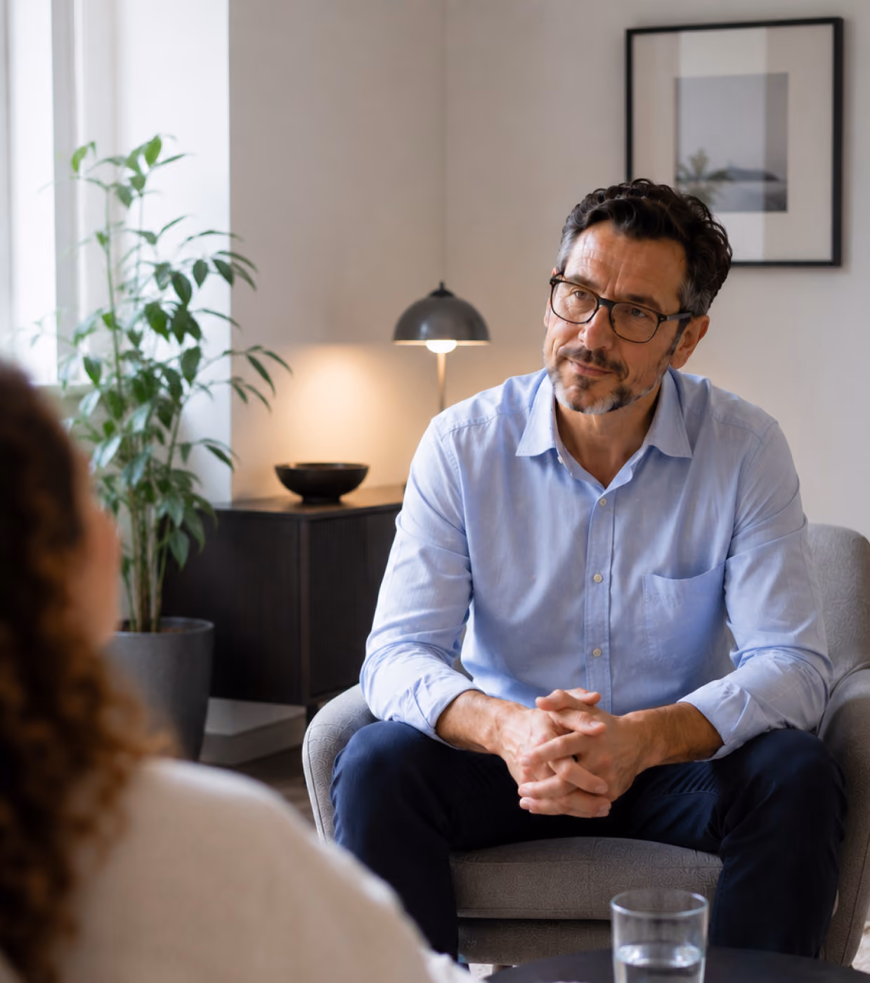 Homme portant des lunettes et une chemise bleu clair, assis dans un fauteuil gris, écoutant attentivement une femme dans une pièce chaleureuse, avec une plante, une lampe et un tableau encadré au mur.