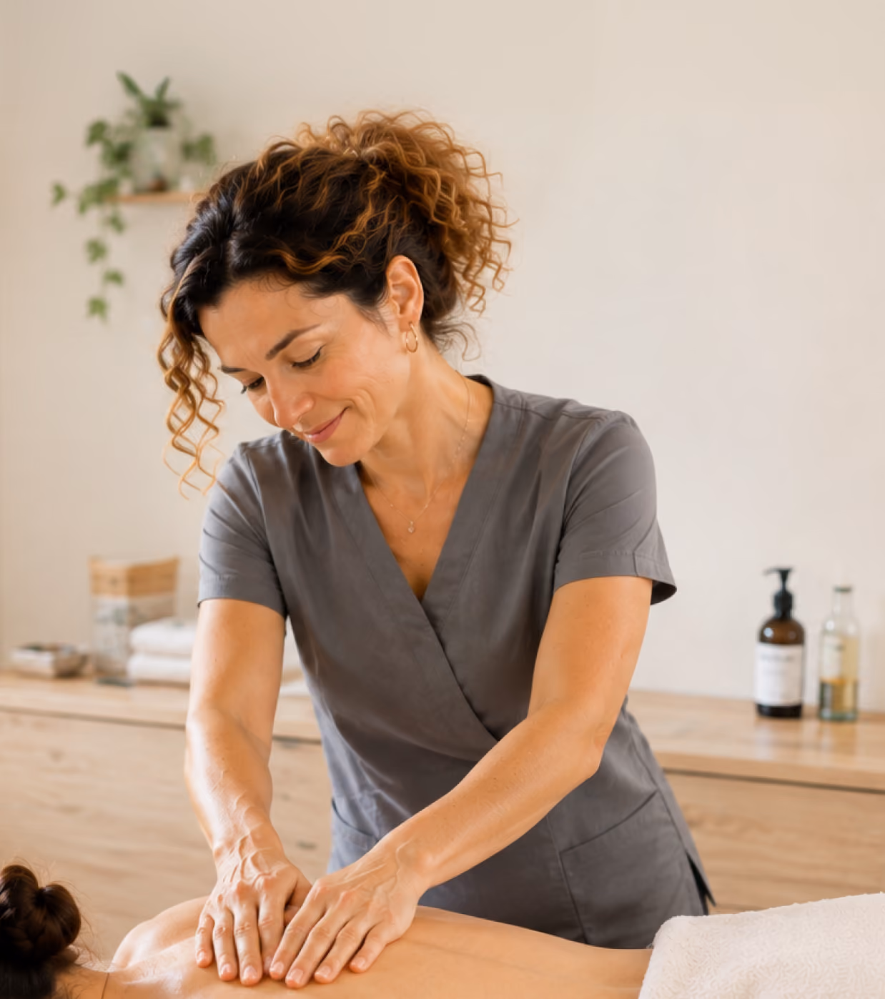 Une masseuse aux cheveux bouclés attachés masse le dos d'une personne allongée sur le ventre sur une table de massage, dans une pièce calme à la lumière tamisée.