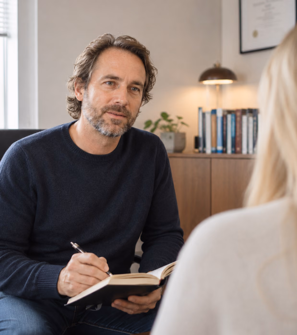 Un homme barbu aux cheveux ondulés, vêtu d'un pull foncé, tient un stylo et un carnet et écoute attentivement une femme blonde vue de dos, dans un bureau chaleureux avec, en arrière-plan, une bibliothèque, une plante et une lampe de bureau.