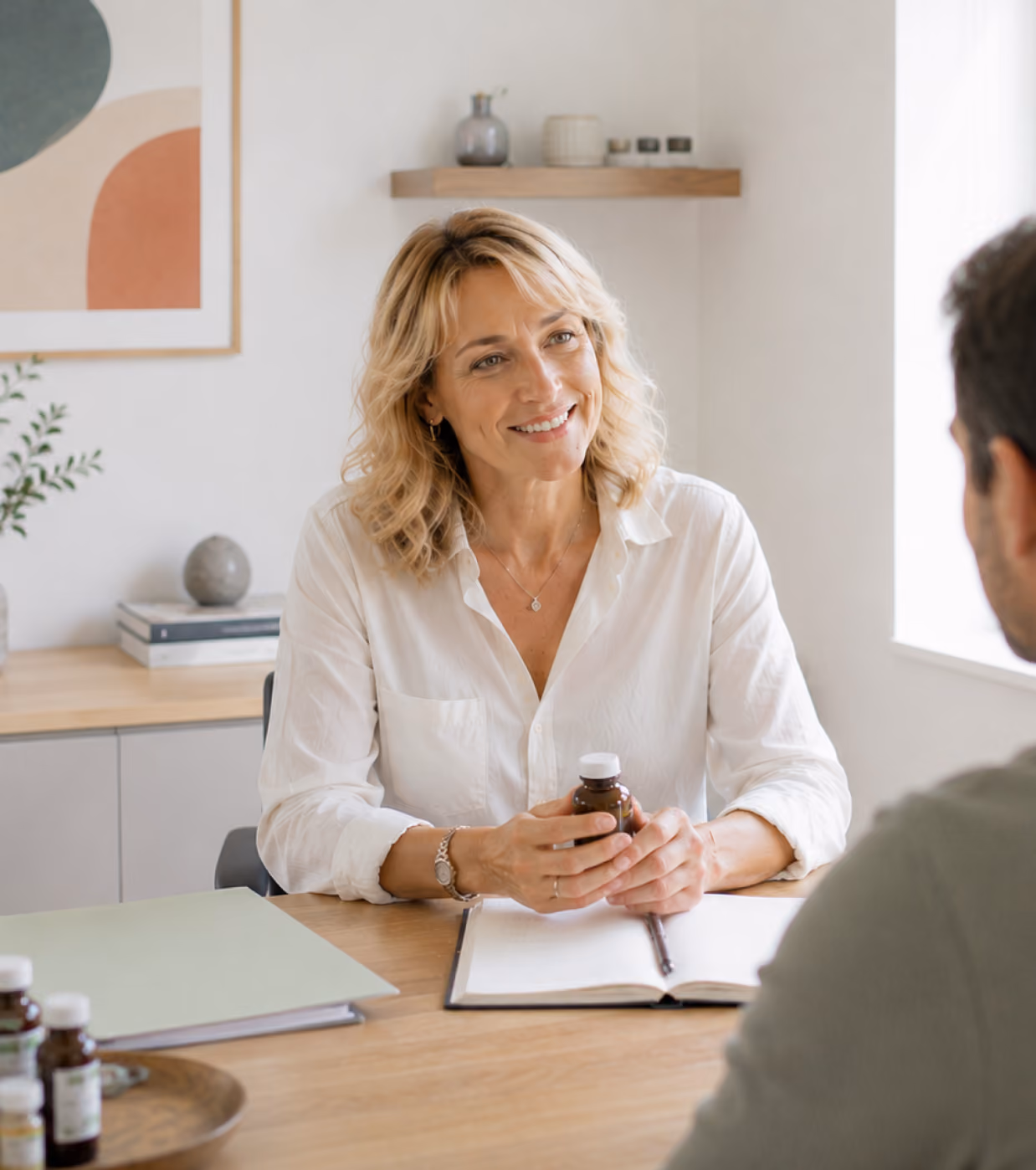 Femme blonde en chemisier blanc, assise à un bureau dans un espace moderne et lumineux, souriante, tenant un petit flacon de médicament tout en parlant à une personne vue partiellement de dos.