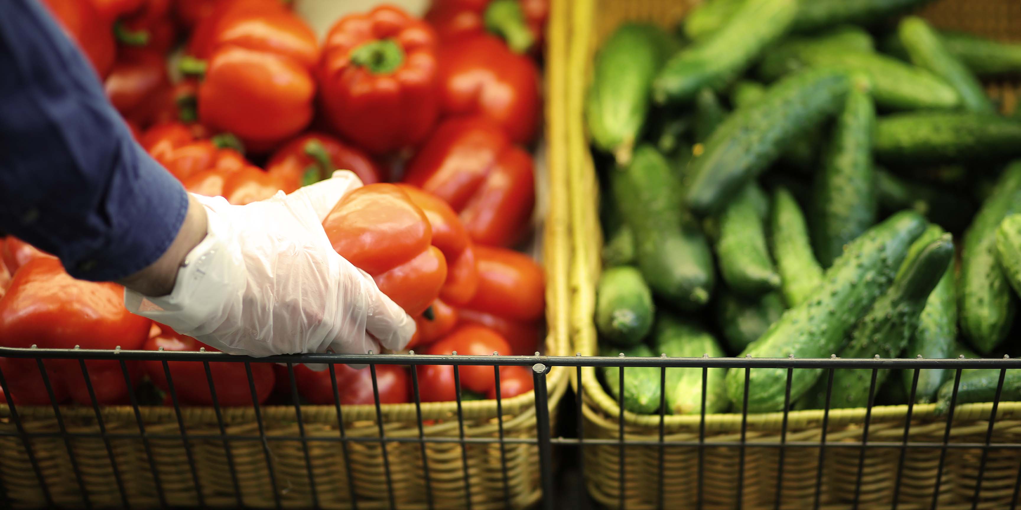 Grocery store worker wearing a glove picking fresh red bell peppers next to cucumbers in baskets