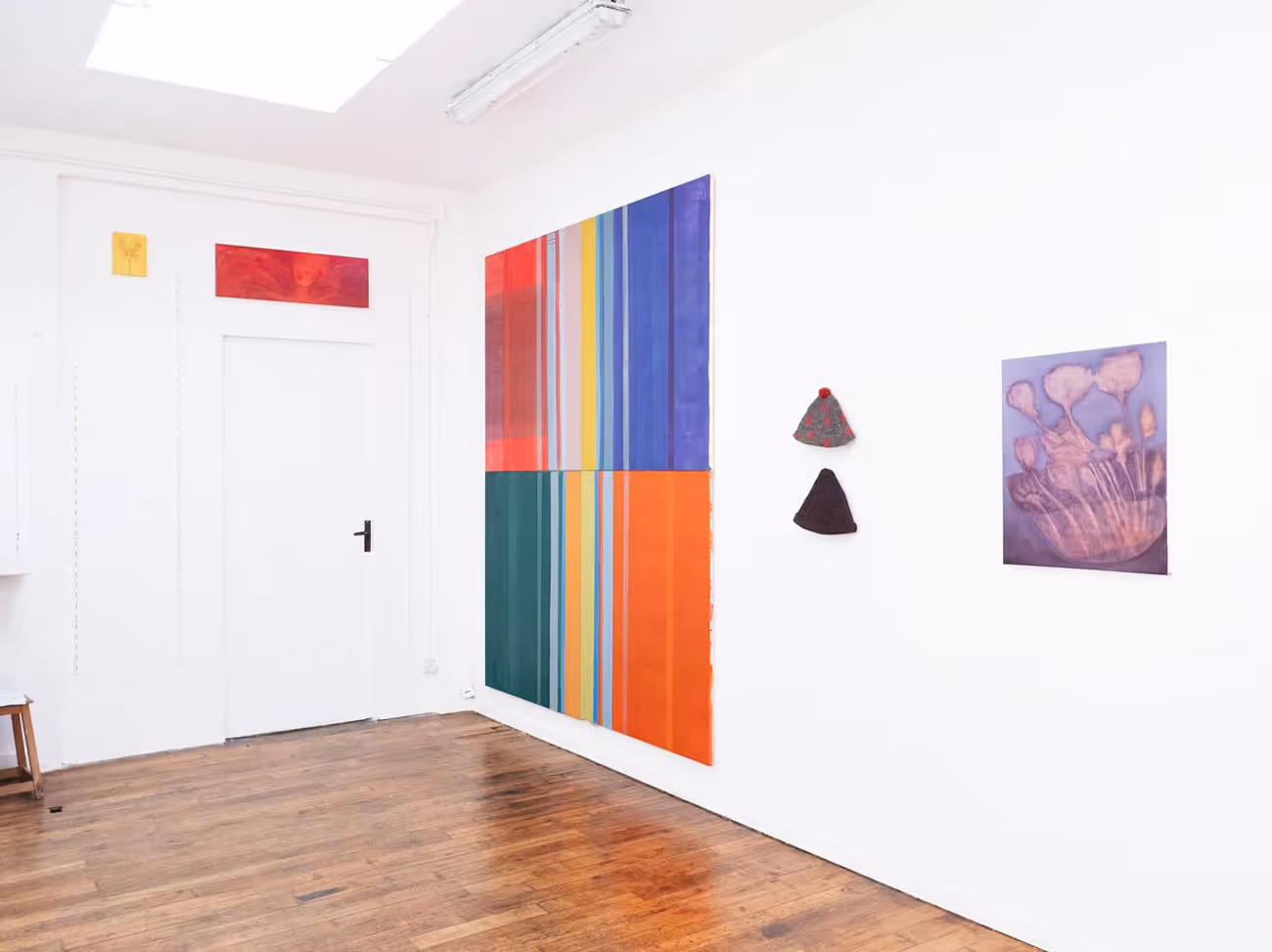 Vue d'une salle d'exposition lumineuse servant aussi d'atelier d'artiste, avec des tableaux colorés et des chapeaux en feutre accrochés sur un mur blanc.