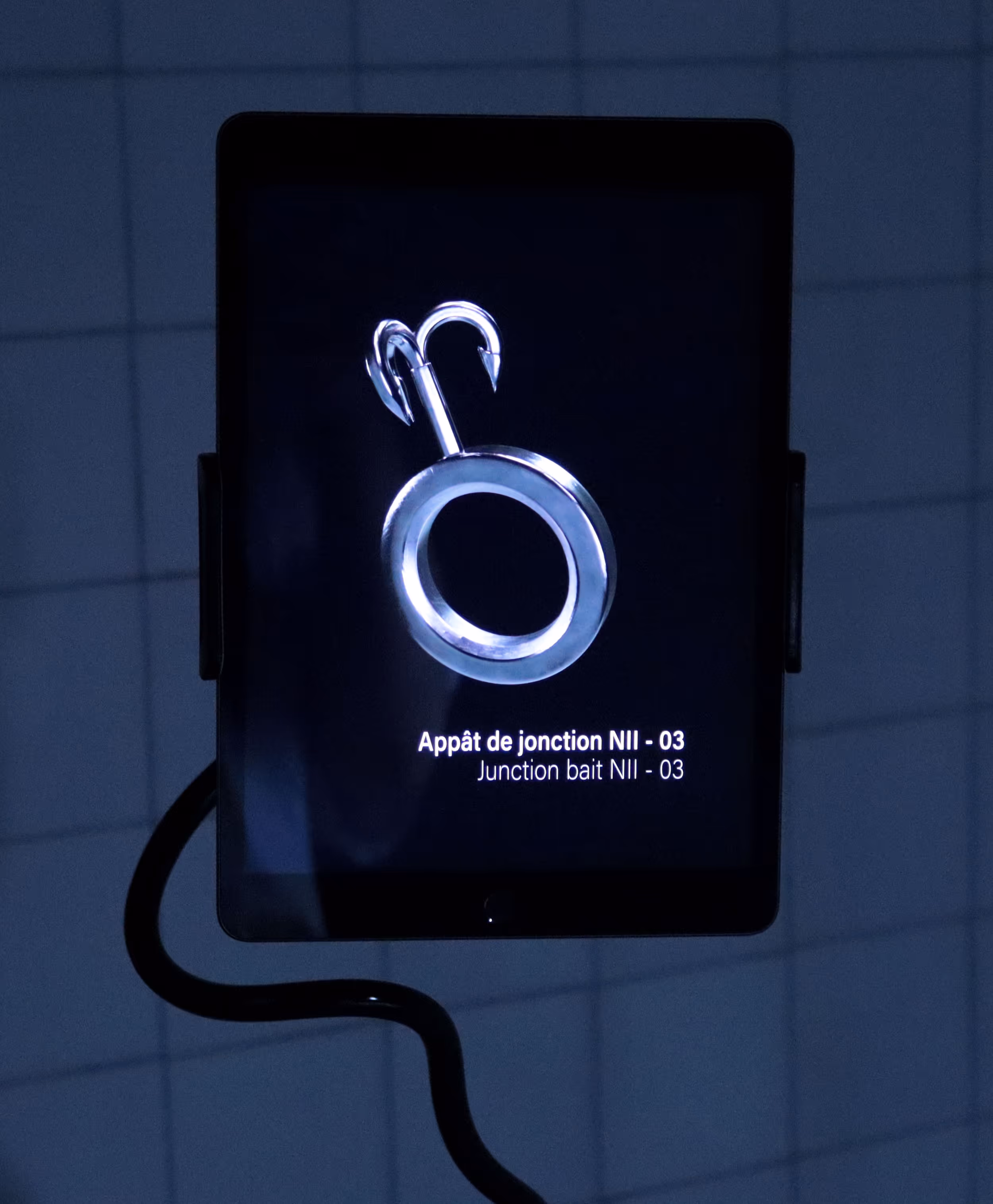 Vue frontale d’une tablette sur laquelle est projeté en 3D un objet hybride : un anneau métallique usé, semblable à un artefact forgé, avec un hameçon de pêche accroché. Cet outil fictionnel, conçu pour collecter le sel, est accompagné de l’inscription : 'appât de jonction N2'.