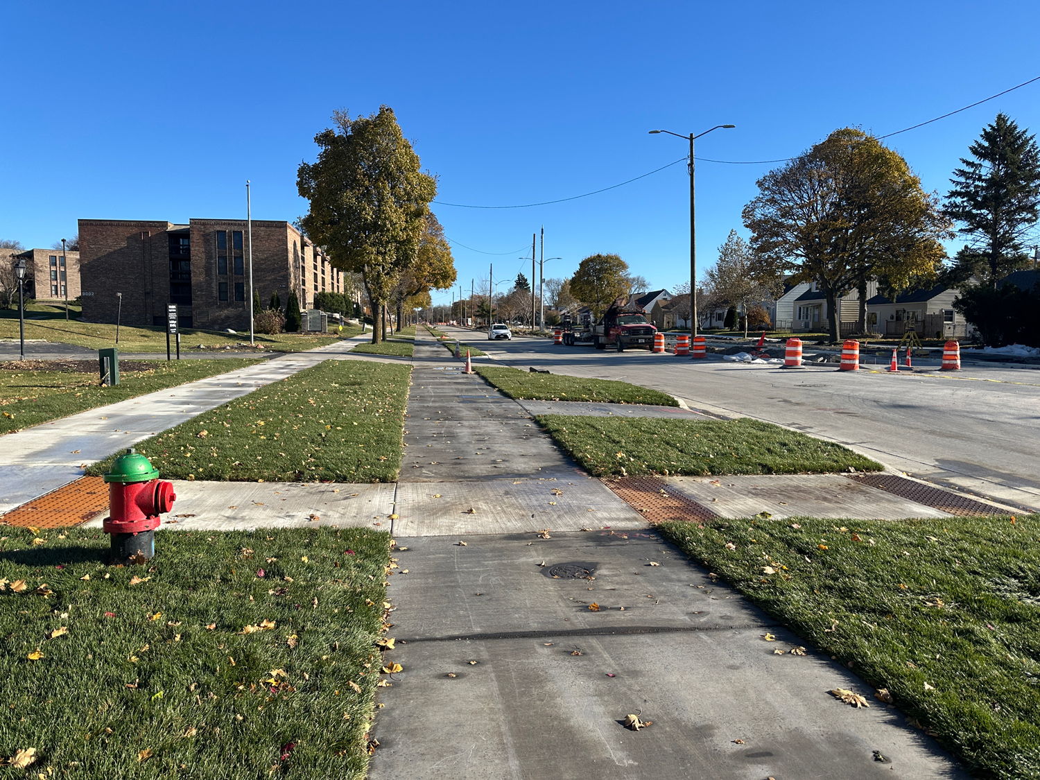Sidewalk and grassy area alongside a street with trees, orange construction barrels, and a red and green fire hydrant under a clear blue sky.