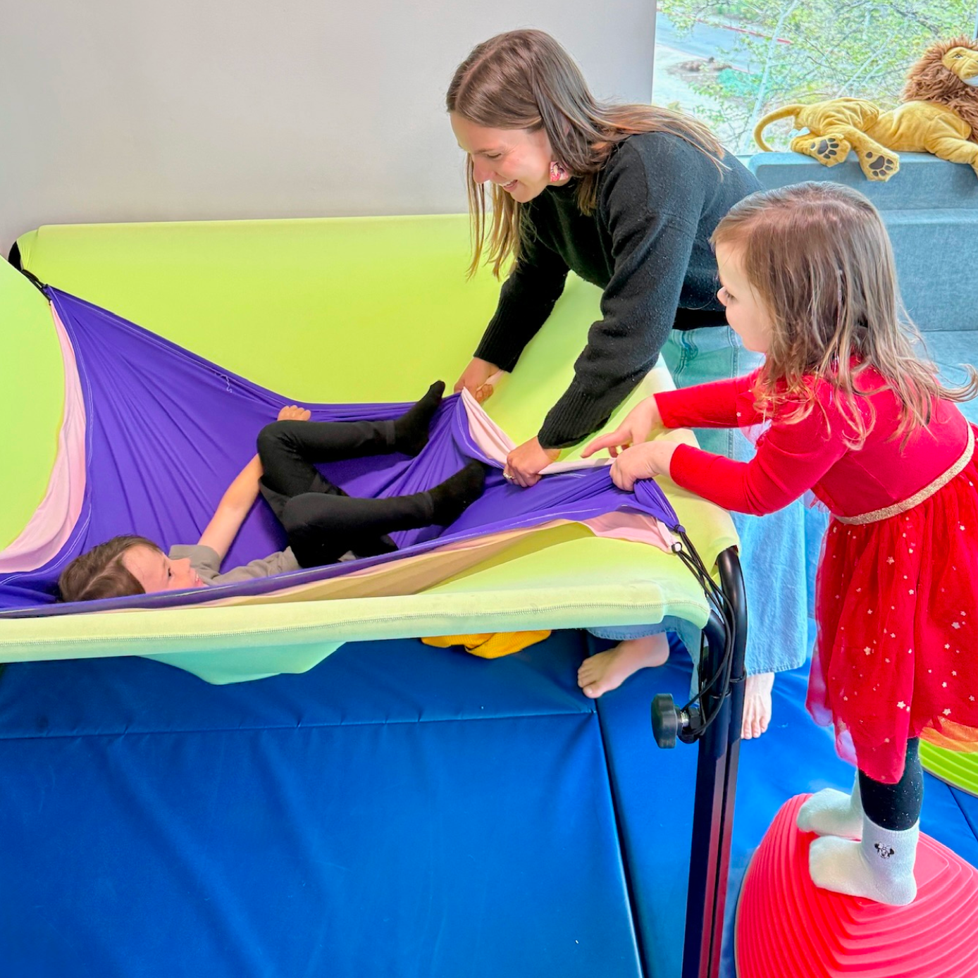 Speech-language pathologist assisting two children using a colorful sensory hammock during a speech therapy session at Speech-N-Motion in San Marcos, CA.