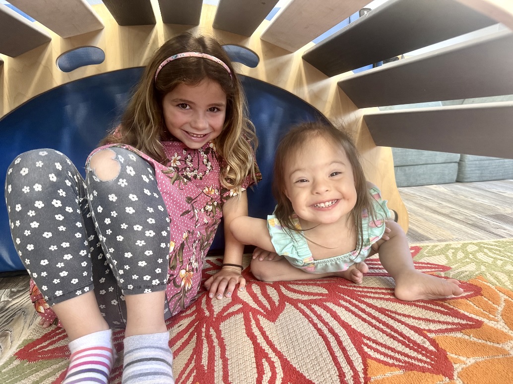 Two young girls smiling while playing under a wooden play structure during a pediatric speech therapy session at Speech-N-Motion in San Marcos, California.