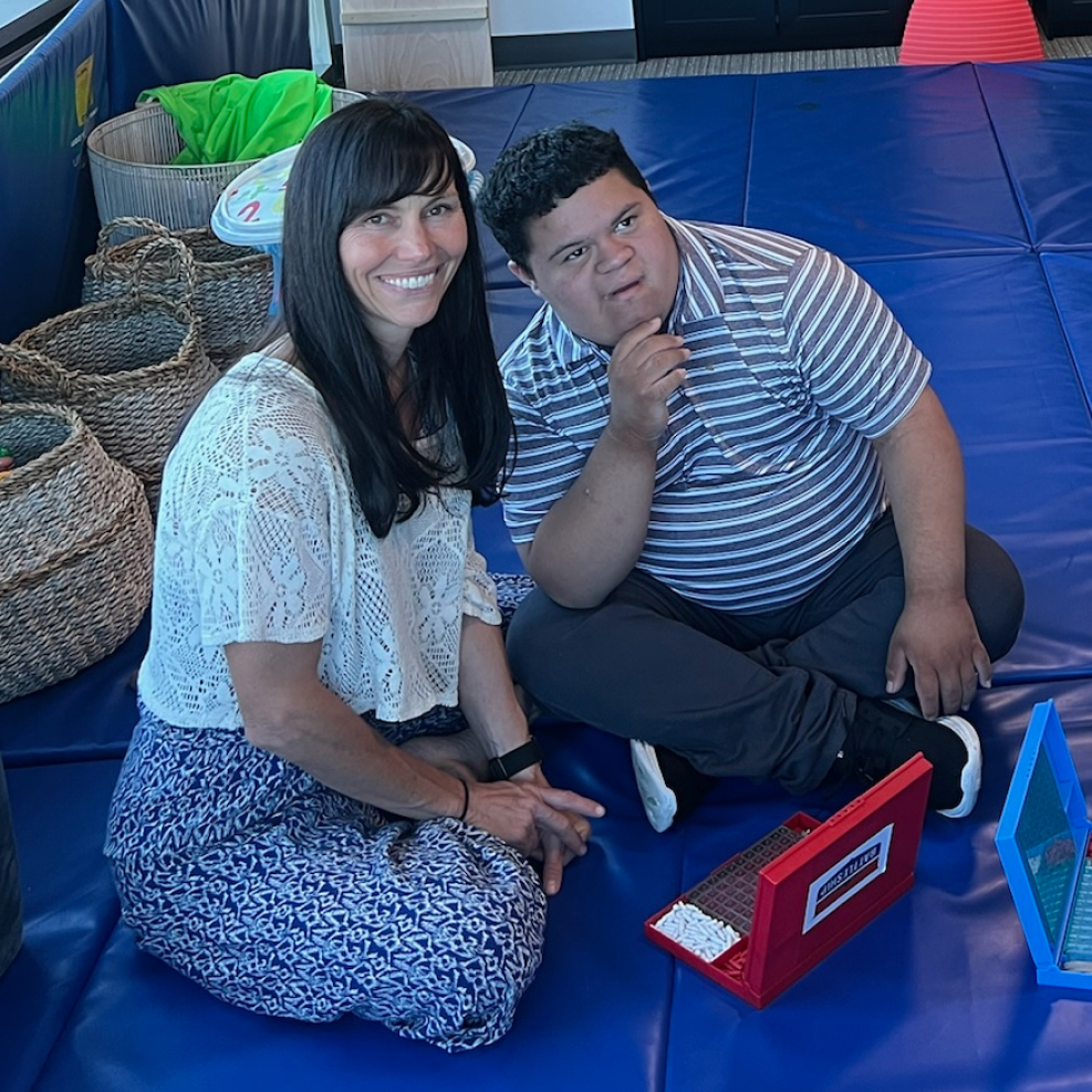 Speech-language pathologist sitting with an older child on a blue therapy mat at Speech-N-Motion, supporting social and communication skills.