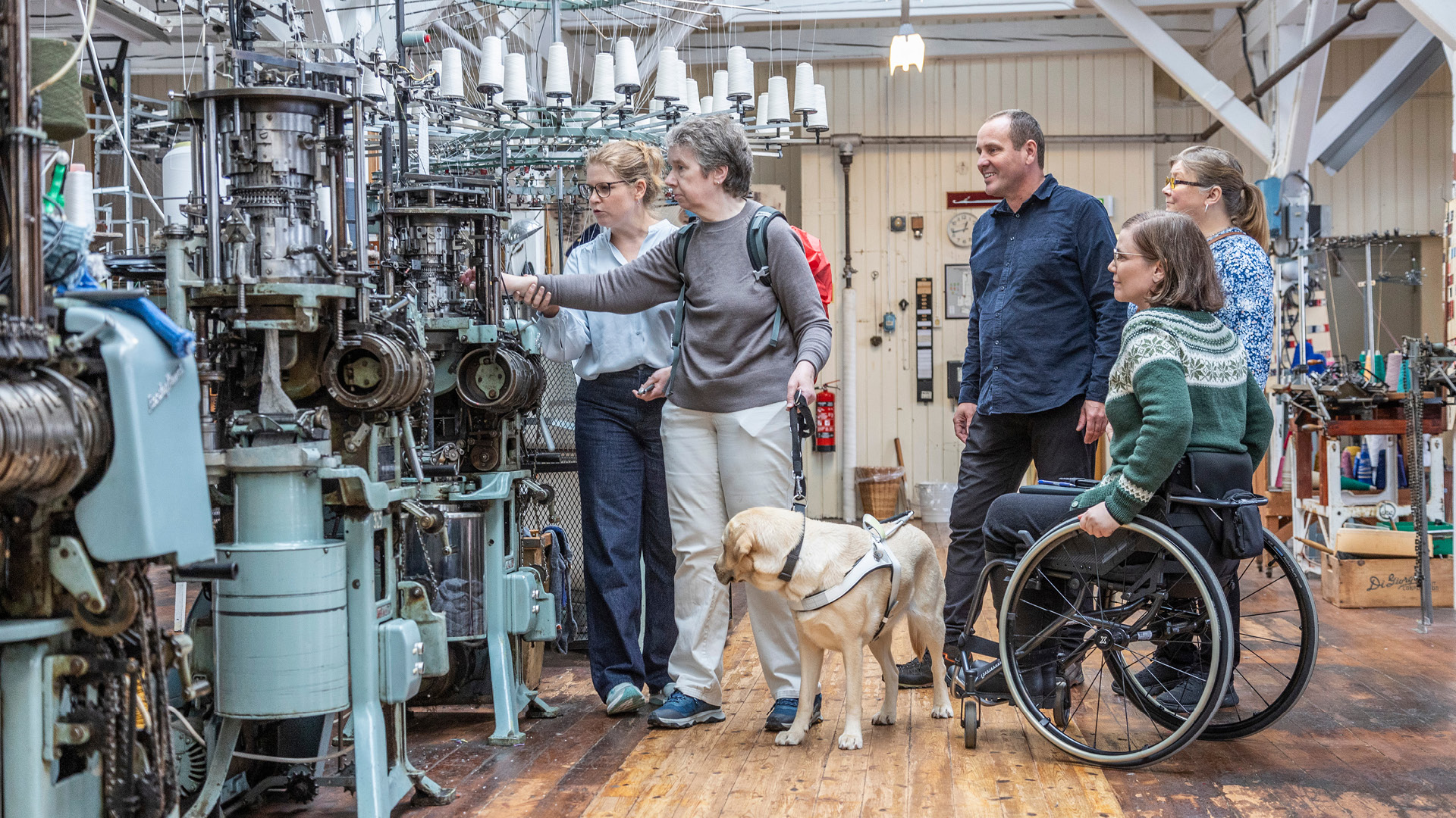 En person med førerhund, en mand i blå skjorte,to kvinder i lyseblå trøjer og en person i kørestol med grøn norsk sweater. De står i et gammelt værksted og rører ved en gammel industri symaskine.