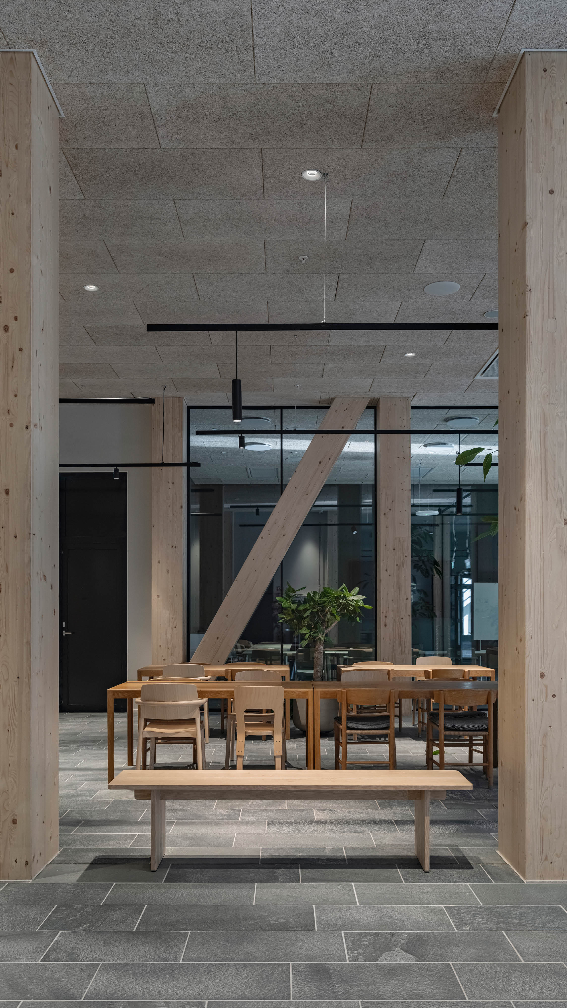 Højformat billede med træsøjler i forgrunden, og længere bord med forskellige træstole rundt om i baggrunden.