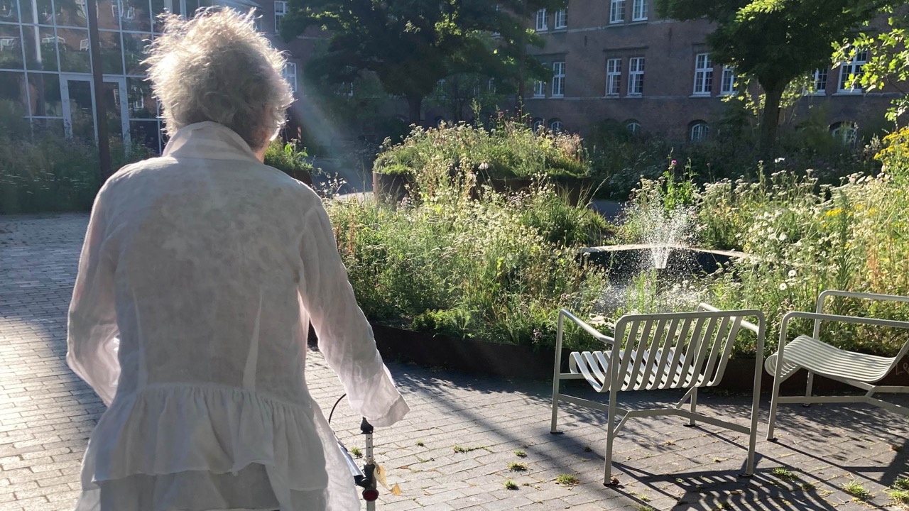 Billede af person med rullator, der går på sti ved siden af stort grønt bed og to grønne havestole