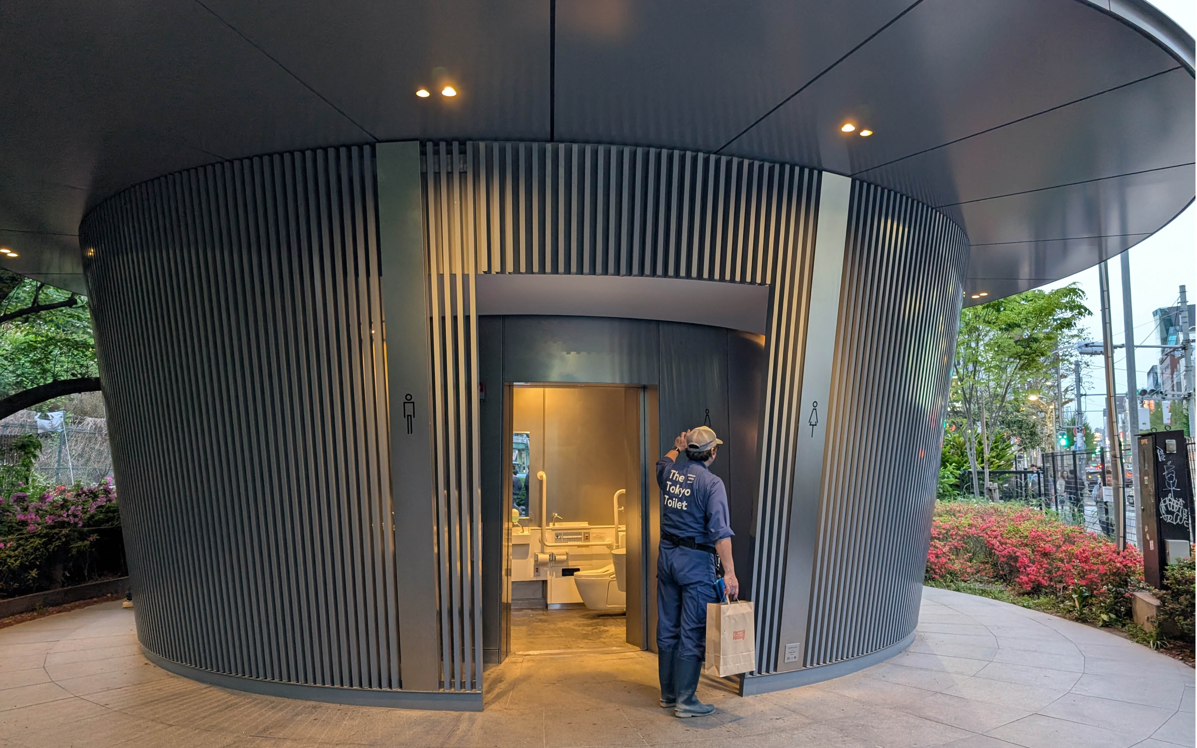 En mand fra Tokyo Toilet er lige blevet færdig med at rengøre et toilet, hvor døren står åbent, så man kan se toiletkumme og håndvask.