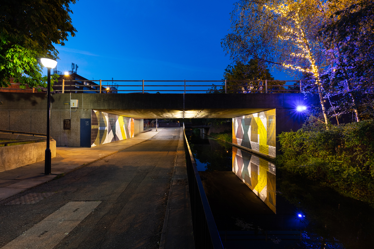 Billede af tunnel om aften, der er oplyst primært på væggene, med flisekunstværk og vandspejl i jorden.