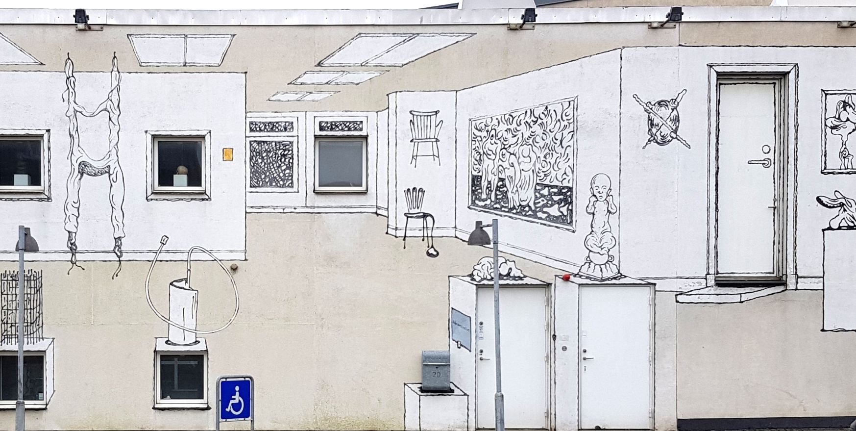 foto af en mur på et muserum, hvor der er tegnet et udstillingsrum med malerier på vægge og skulpturer på gulvet. Foran ser man et parkeringsskilt ift. handicapegnede biler. 