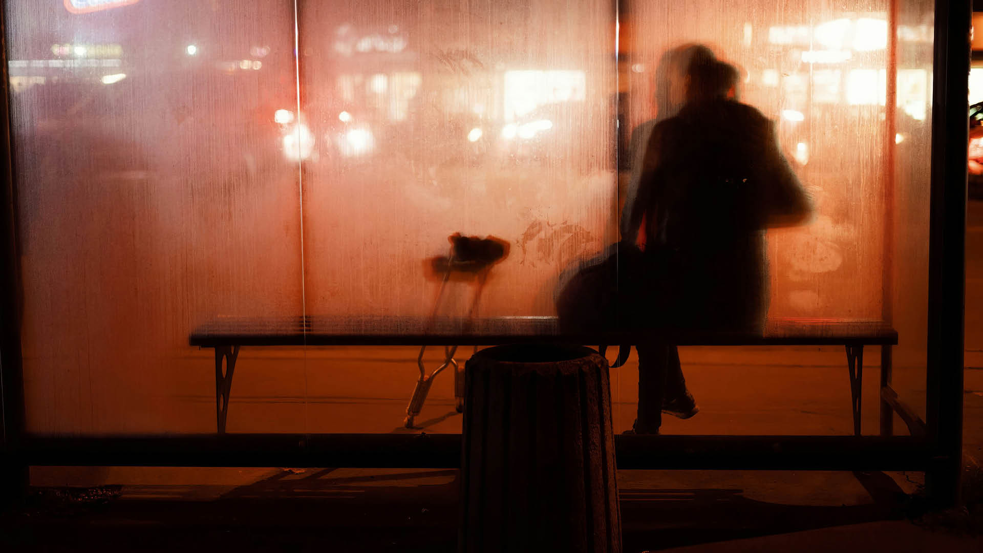 Person sidder på en bænk bag matteret glas ved et busstoppested om aftenen, med et par krykker lænet op ad bænken.