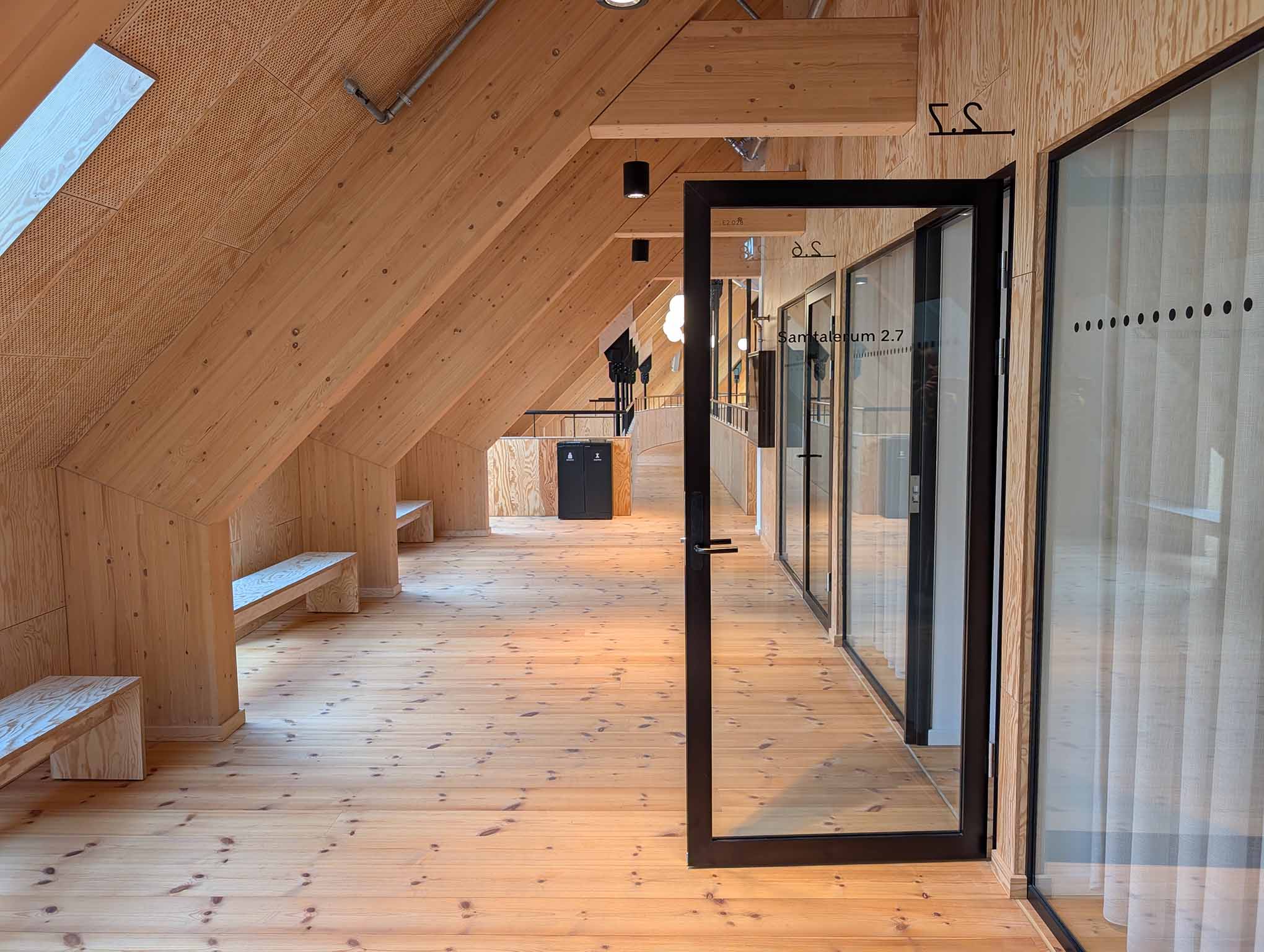 Gangareal med skråt loft, indbyggede bænke og adgang til mindre samtalerum med glaspartier.