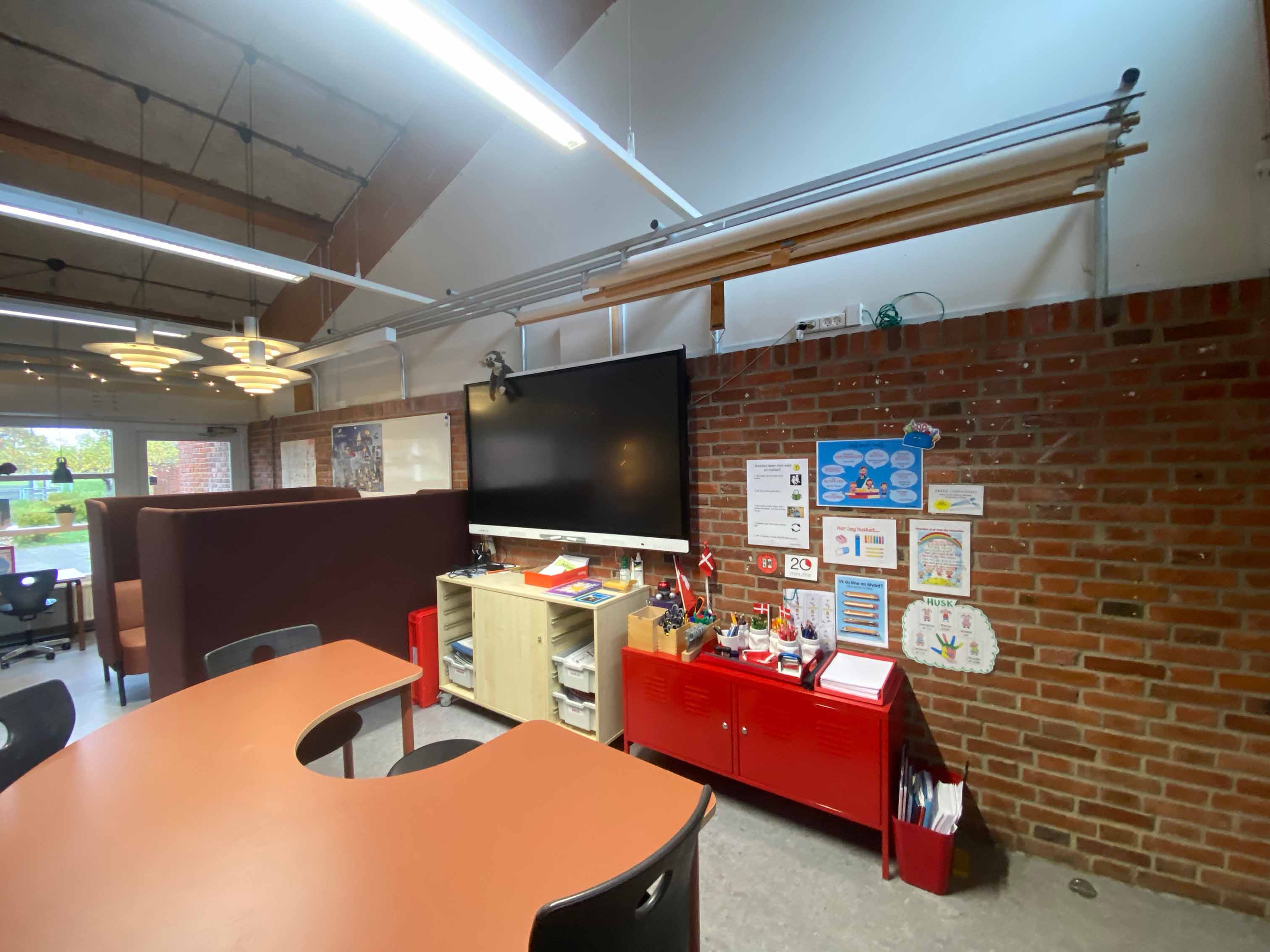 Klasserum med murstensvæg, skærm, opbevaringsmøbler, et halvmåneformet bord og to sofaer i baggrunden med høj ryg. 