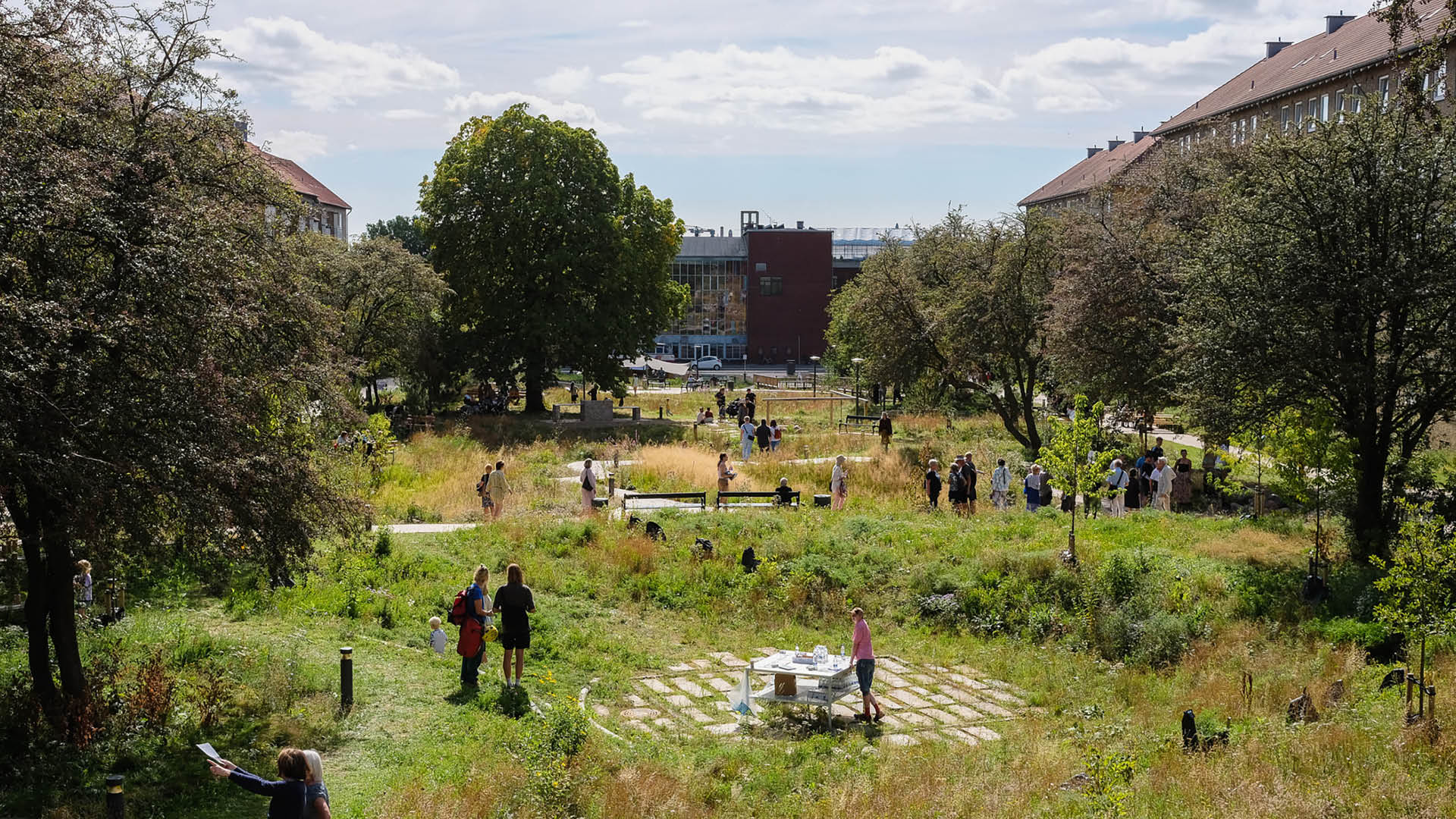 Et grønt byrum med planter, stier og opholdszoner, hvor mennesker går, opholder sig og mødes i landskabet mellem boligbygninger.