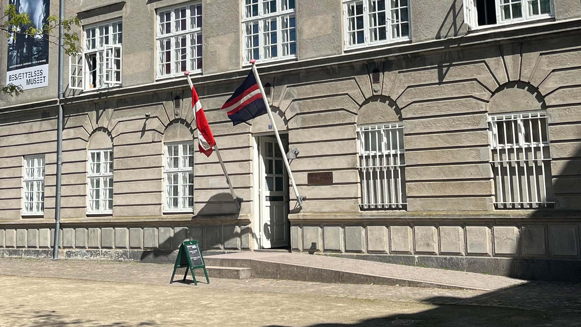 Klassisk bygning med flag ved indgangen, en rampe og en trappe op til døren samt et skilt foran på en solrig forplads.