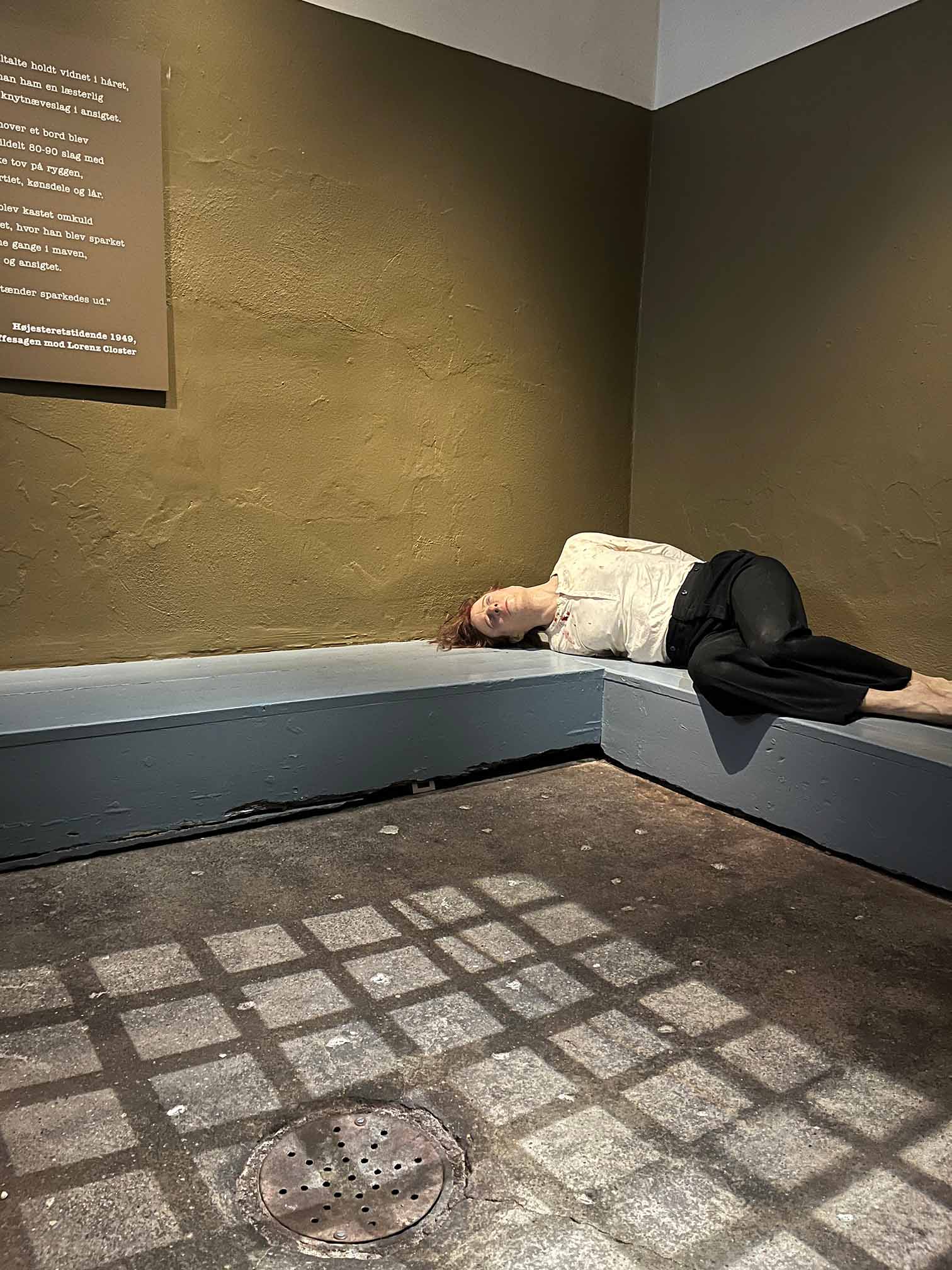 En dukke ligger udstrakt på en bænk i et råt rum med skygger fra tremmer på gulvet.