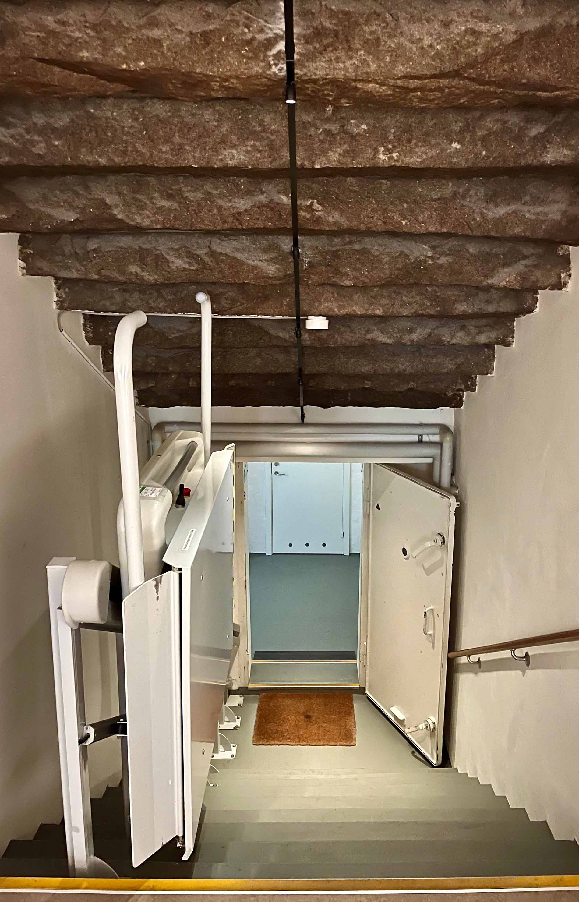 En smal trappe med en stolelift fører ned til en åben dør i et kælderlignende område.