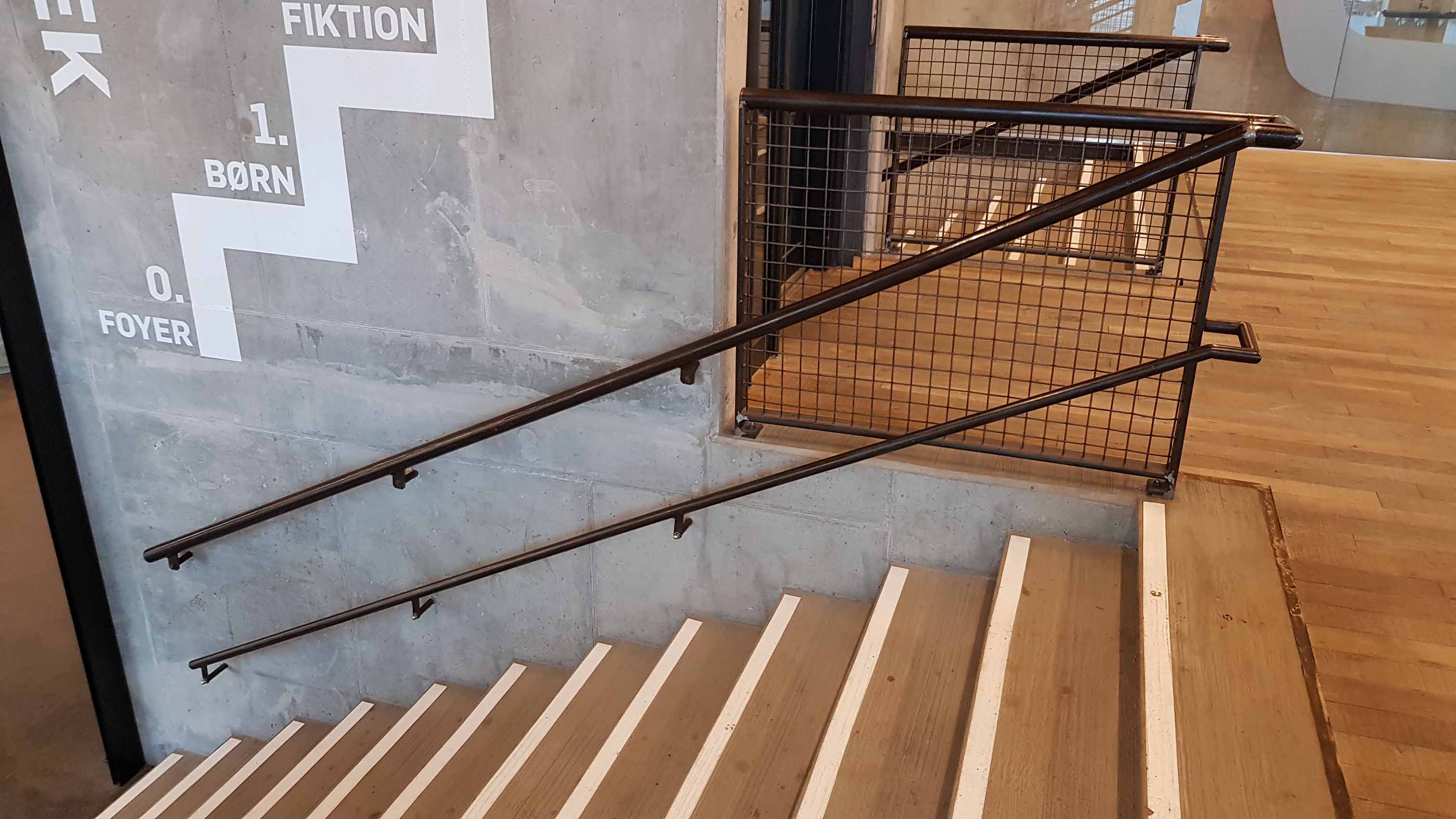 Trappe i lyst træ med hvide kontrastmarkeringer på trinene, dobbelte håndlister og betonvæg med wayfinding-grafik.