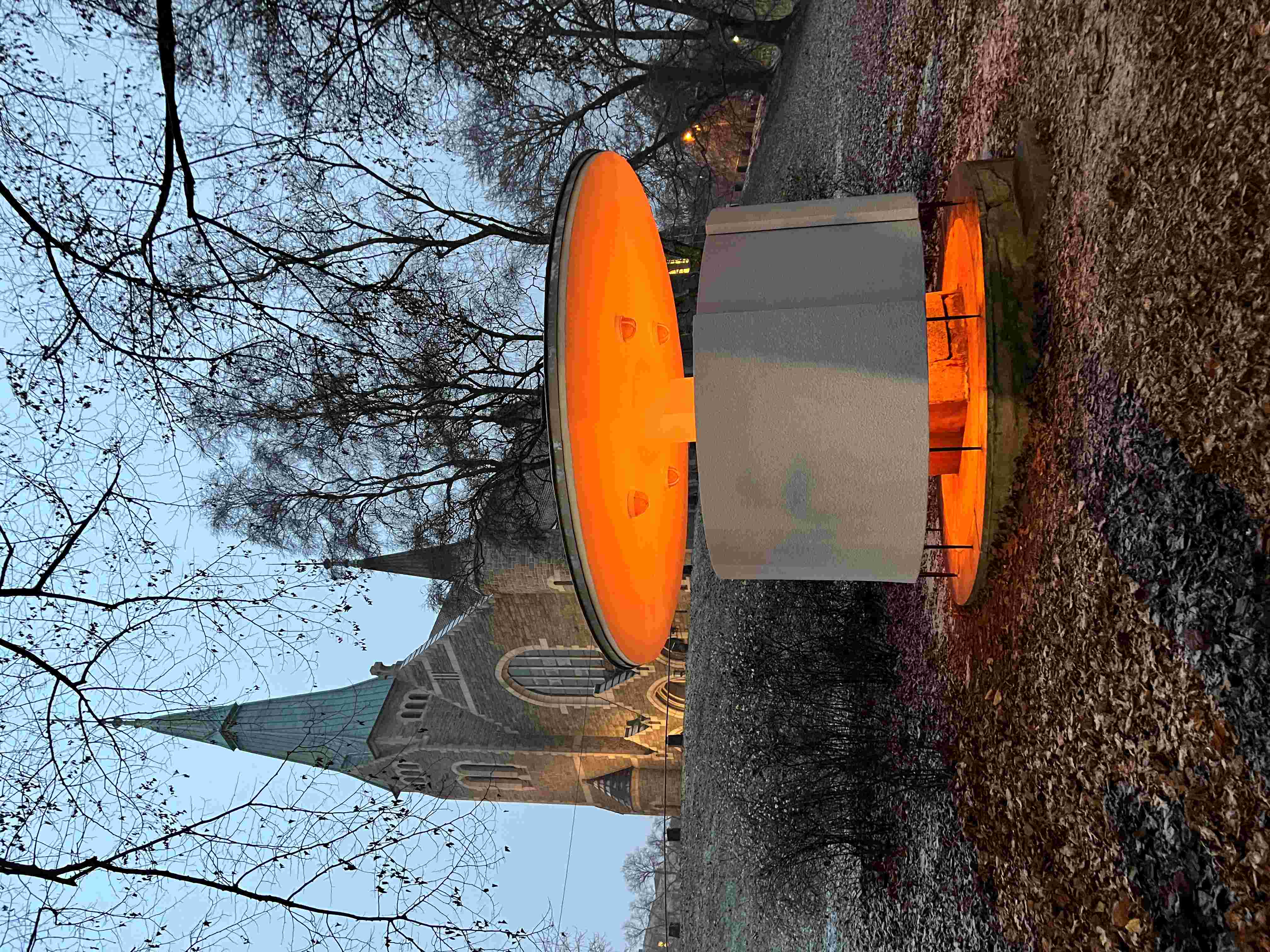 Aftenfoto af et pissoir i beton med et rundt tag. Varmt lys strømmer ud. I baggrunden en kirke. 