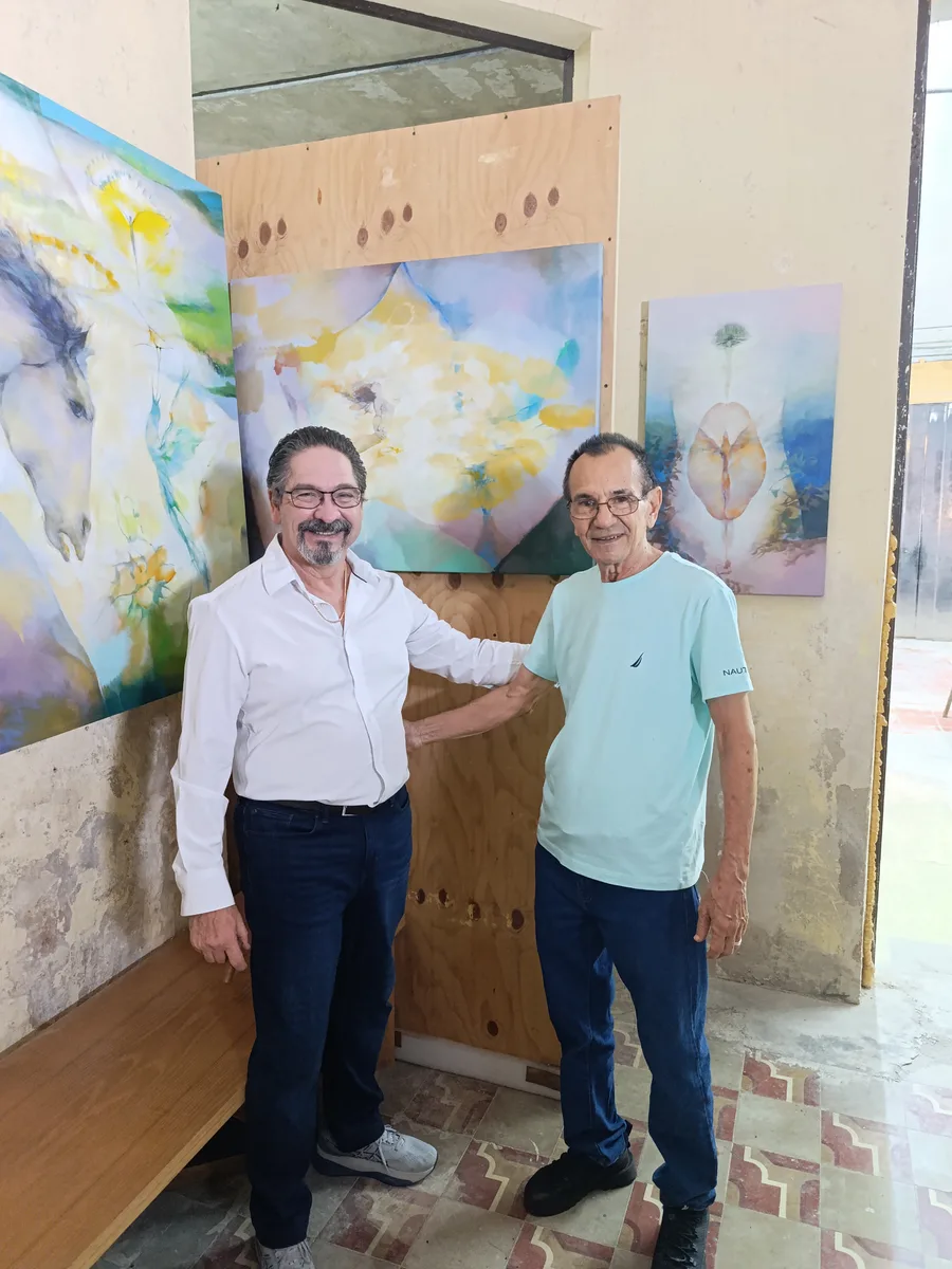 Two men posing together in front of colorful abstract paintings in the studio.
