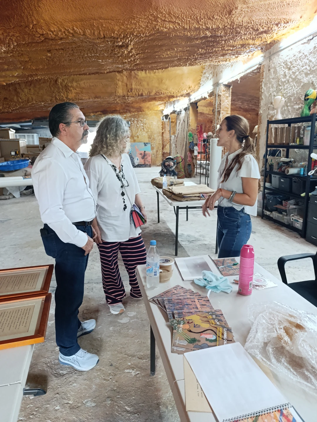 Leonardo Rodríguez, Claribel Calderius and Ivonne Ferrer talking in the artist's studio, surrounded by work tables and art materials.