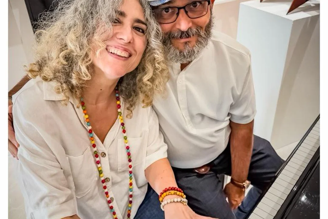 Ivonne Ferrer and Gary A. Añuez smiling together at a piano in her Florida studio, with artworks on the wall behind them