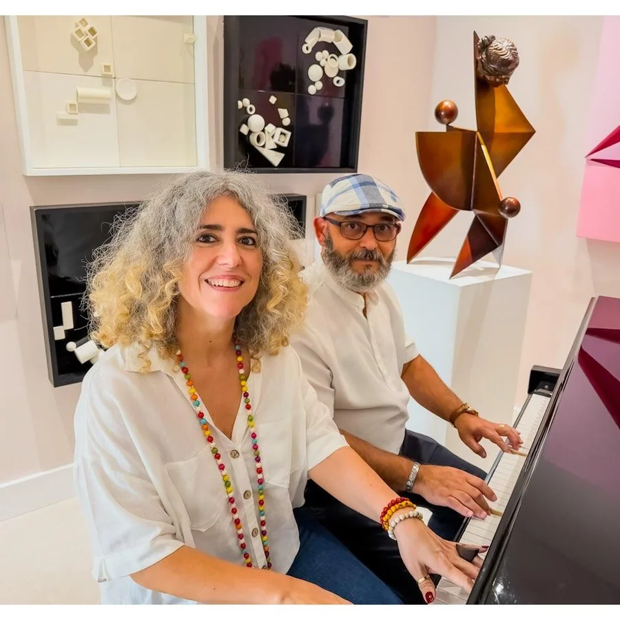 Ivonne Ferrer and Gary A. Añuez with their hands on the piano keys in her studio, geometric reliefs and a bronze sculpture behind them