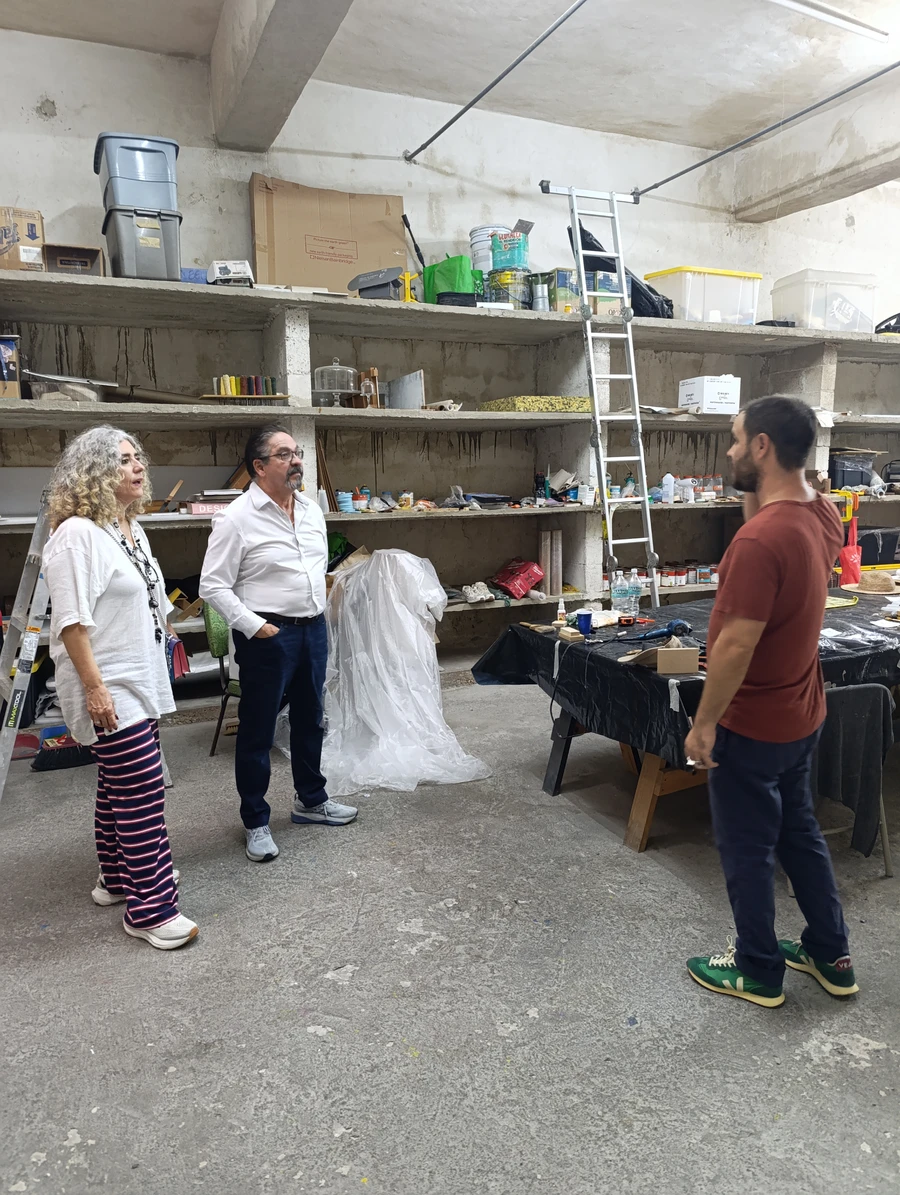 Three people talking inside the artist's studio, among shelves with art materials, a ladder and a work table.