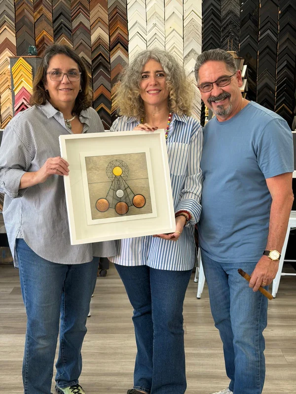 Three people holding the framed artwork by Rosario Verdecia in front of a wall of frame samples