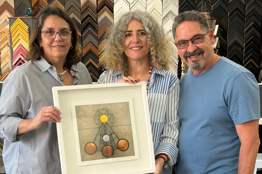 Three people holding the framed artwork by Rosario Verdecia in front of a wall of frame samples