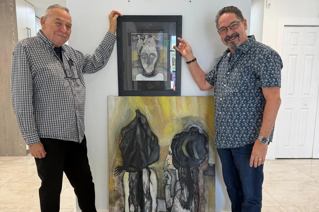 Two men standing beside two works by Alejandro Lorenzo Valdés (ALEVAL), a framed drawing and a painting.