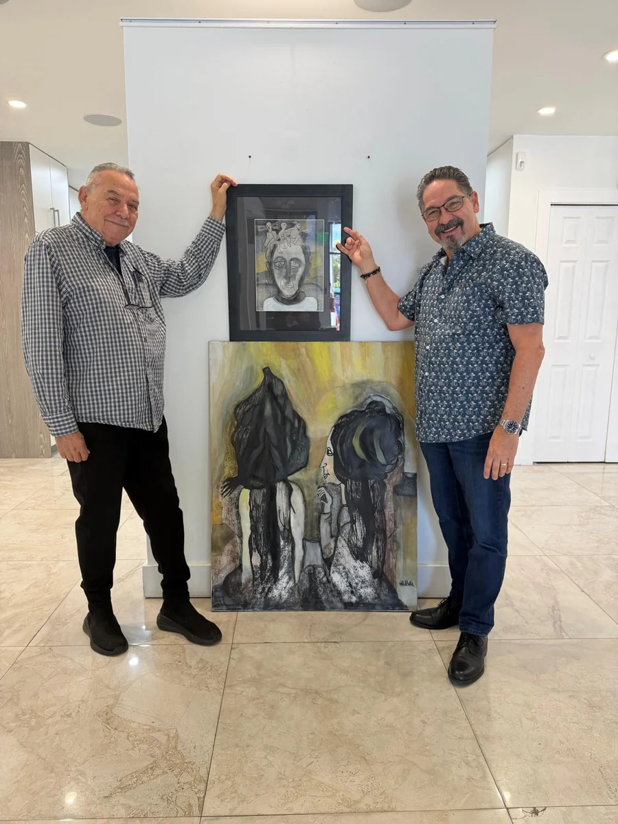 Two men standing beside two works by Alejandro Lorenzo Valdés (ALEVAL), a framed drawing and a painting.