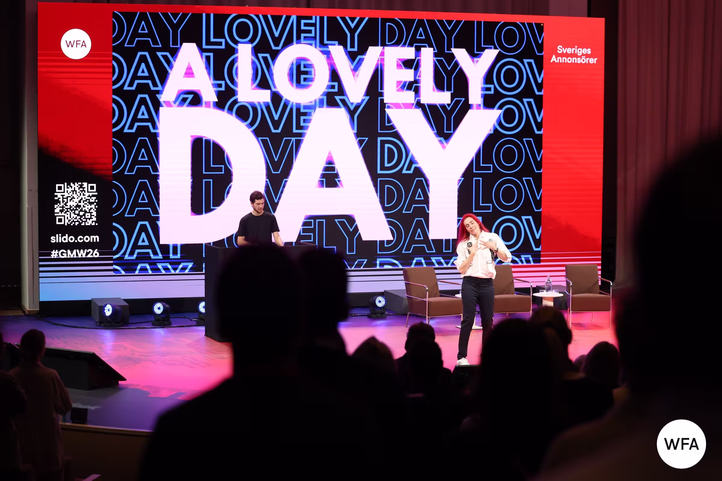 A speaker with red hair and a white jacket addresses the audience on stage at a WFA Global Marketer Week event, with a large LED screen behind her displaying the words "A Lovely Day" in bold white lettering.